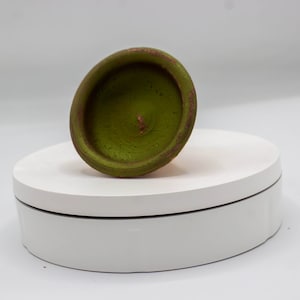 Op de afbeelding: Een kleine, groene keramische kom met een roodbruine rand, gekanteld op een wit draaiend platform. De binnenkant van de kom is een lichtere tint groen en de rand vertoont tekenen van slijtage. De achtergrond is effen wit.