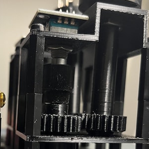 Puede incluir: Primer plano de un mecanismo de engranaje impreso en 3D negro con una pequeña placa de circuito y una perilla negra. El mecanismo de engranaje está montado en una base negra.