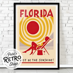May include: A framed vintage travel poster for Florida. The poster features a red sun, a woman in a lounge chair, and the words "FLORIDA" and "MUST BE THE SUNSHINE!" in red. The background is a cream color with yellow and tan accents.