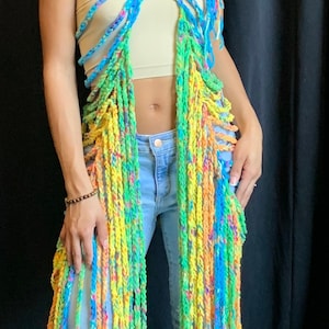 May include: A colorful, handmade, open-front garment with long, fringed strands. The piece is crafted from yarn in shades of blue, yellow, green, and orange. The model is wearing a beige tube top and blue jeans.