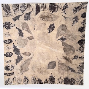 May include: A beige textile with a repeating pattern of leaf imprints in shades of gray and black. The leaves vary in size and shape, creating a natural, organic design. The edges are finished with a stitched border.
