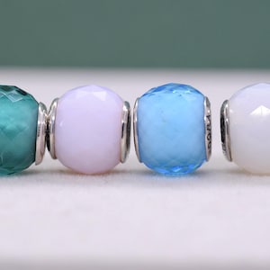 Puede incluir: Cuatro cuentas de vidrio facetadas en verde azulado, lavanda, azul claro y blanco. Cada cuenta es redonda con una banda de metal plateado. Las cuentas son probablemente para una pulsera o collar.