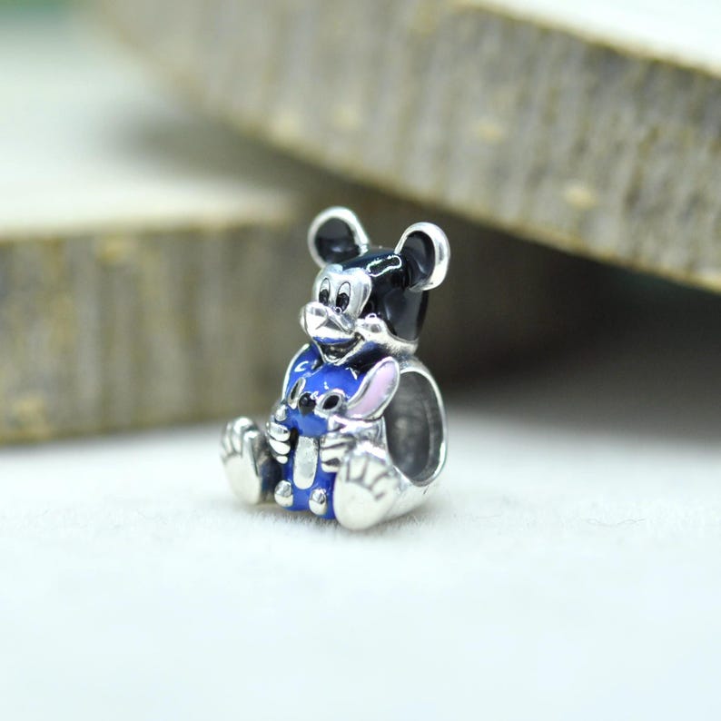 Puede incluir: Un abalorio de plata que muestra una figura de Mickey Mouse en blanco y negro sosteniendo a un peque&ntilde;o personaje de Stitch azul y rosa. El abalorio tiene un centro hueco para pulsera, con coloraci&oacute;n esmaltada detallada y un acabado met&aacute;lico pulido.
