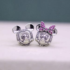May include: A pair of silver-toned Mickey and Minnie Mouse stud earrings. Mickey has a round face with a smiling expression, and Minnie has a pink bow. Both are embellished with small, clear crystals around the face.