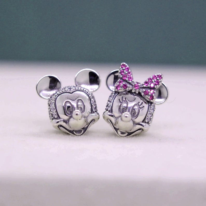May include: A pair of silver-toned Mickey and Minnie Mouse stud earrings. Mickey has a round face with a smiling expression, and Minnie has a pink bow. Both are embellished with small, clear crystals around the face.