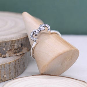 Op de afbeelding: Een zilveren ring met een hartvormig ontwerp. De ring heeft een rij kleine, heldere edelstenen langs de bovenkant van het hart. De ring wordt gepresenteerd op een houten kegelvormige standaard.