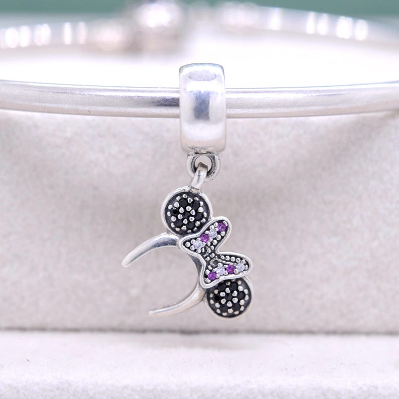 May include: A silver charm with a butterfly and two black dotted circles. The butterfly has purple and white stones. The charm is attached to a silver bracelet.