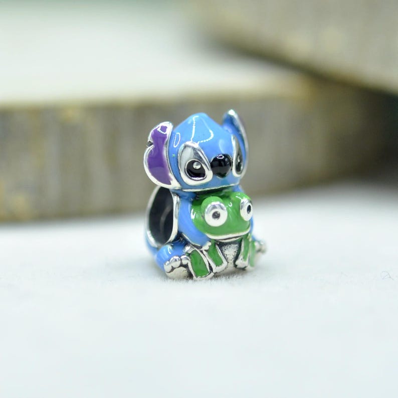 May include: A silver charm bead featuring a blue character with large ears and purple inner ear details, holding a small green frog. The charm has enamel accents in blue, purple, and green, with a hollow center for a bracelet or necklace chain.