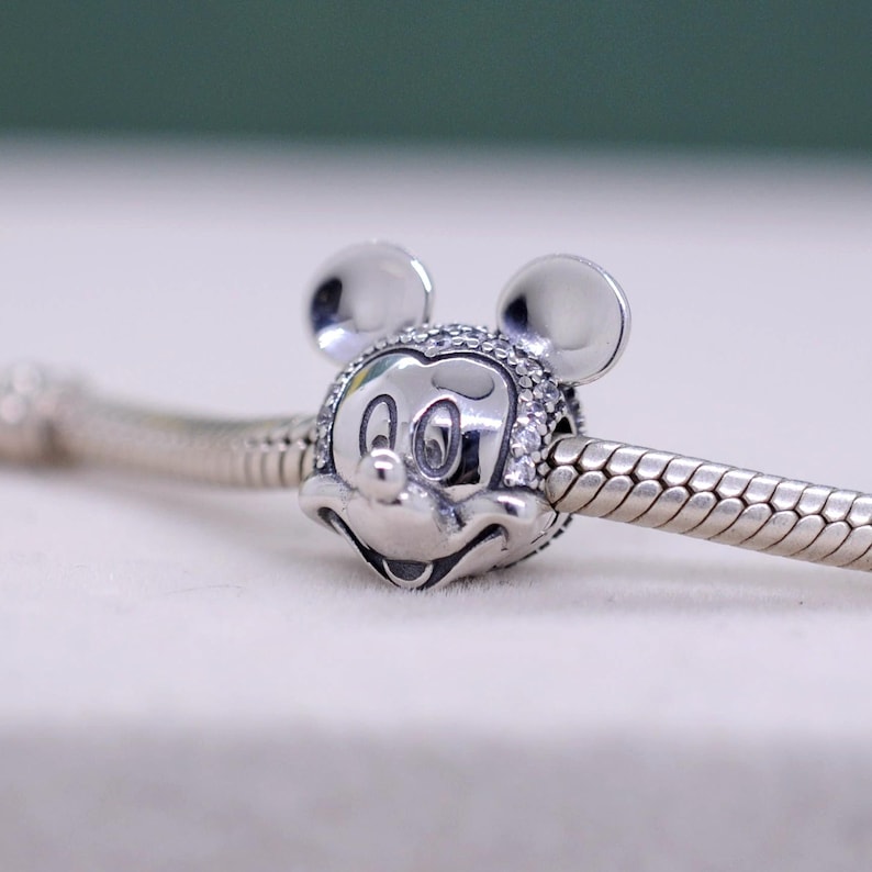 May include: A silver charm featuring Mickey Mouse's face, with his iconic ears and a smiling expression. The charm is adorned with small, clear stones around the face and is strung on a silver chain. The charm is a Disney-themed jewelry item.