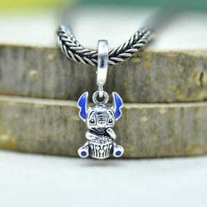 May include: A silver charm pendant featuring a cartoon character with blue ears and accents. The charm is suspended from a braided silver chain. The character is holding a cupcake. The charm is approximately 1 inch in height.