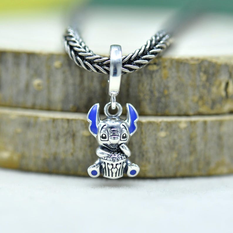 May include: A silver charm pendant featuring a cartoon character with blue ears and accents. The charm is suspended from a braided silver chain. The character is holding a cupcake. The charm is approximately 1 inch in height.