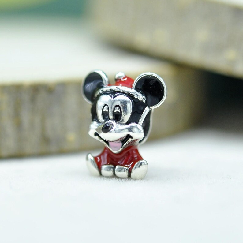 May include: A silver-toned metal charm featuring a character with large round black ears, wearing a red Santa hat with white trim and a matching red outfit. The character has a smiling face with a black nose and a small pink tongue visible.