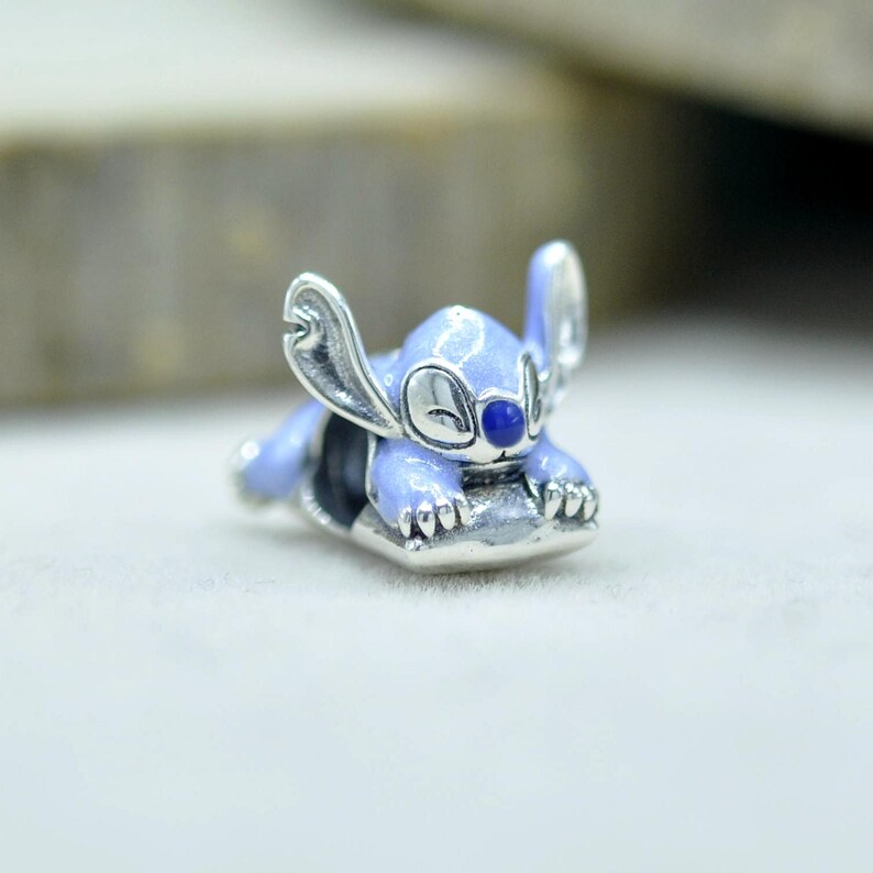May include: A silver charm bead featuring a light blue enameled character with large ears and a dark blue nose, depicted in a resting position. The metal surface is polished and reflective, showcasing detailed engravings on the ears and paws.