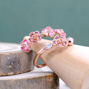 Może przedstawiać: Pierścionek z różowego złota z kwiatowym wzorem z różowymi emaliowanymi kwiatami i małymi, okrągłymi, różowymi kamieniami szlachetnymi. Pojedynczy, przezroczysty kamień szlachetny jest osadzony na obrączce. Pierścionek jest prezentowany na drewnianym kołku na turkusowym tle.