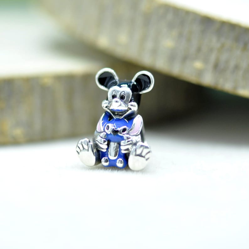 Puede incluir: Un dije de metal plateado que muestra un personaje de rat&oacute;n en blanco y negro sentado, sosteniendo una peque&ntilde;a criatura azul y rosa. El dije tiene detalles en plata pulida en las orejas, cara, manos y pies, con detalles en esmalte negro y azul.