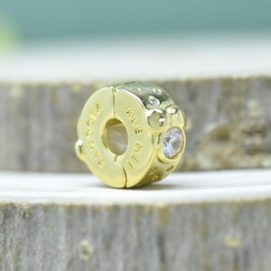 Puede incluir: Un charm dorado con el texto grabado "PANDORA ALE MET" y peque&ntilde;as piedras preciosas transparentes. El charm es redondo con un dise&ntilde;o de cierre, sobre un fondo neutro.