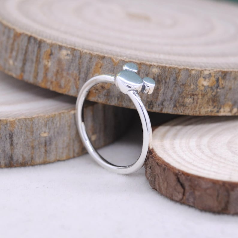 May include: A silver ring featuring a three-circle design, reminiscent of a famous cartoon character. The ring is set against a backdrop of wooden slices, highlighting its simple yet recognizable form. The ring's design is a playful and whimsical accessory.
