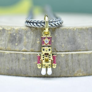 May include: A gold-colored nutcracker charm with red and white accents. The charm features a red hat with a star and a clear gem. The arms and details are red, and the feet are white. The charm is attached to a silver chain.