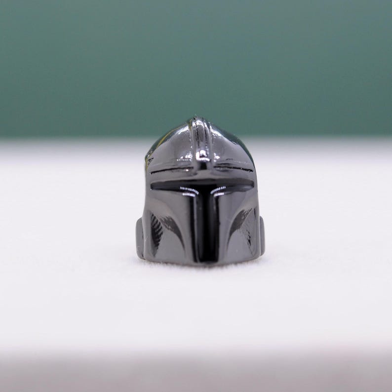 Puede incluir: Un peque&ntilde;o casco gris met&aacute;lico con una visera en forma de T y una cresta distintiva. El dise&ntilde;o del casco recuerda a un casco mandaloriano, con un acabado liso y pulido. Es un art&iacute;culo de colecci&oacute;n.