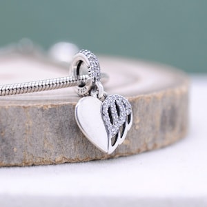 May include: A silver charm featuring a heart and wing motif. The heart is divided, with one half plain and the other embellished with tiny, glittering stones. The charm is attached to a silver bracelet. The charm is marked with "S925" and "ALE".
