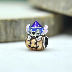 May include: A silver-toned metal charm bead featuring a blue character with large ears wearing a purple pointed hat, emerging from a tan-colored jack-o'-lantern pumpkin with a carved face. The bead has a hollow center for a bracelet chain.