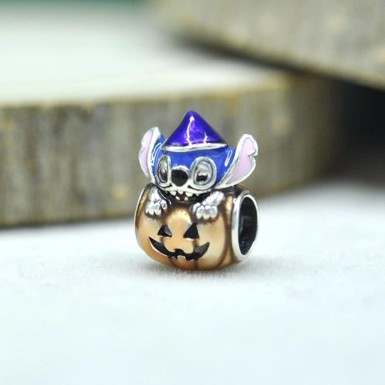 May include: A silver-toned metal charm bead featuring a blue character with large ears wearing a purple pointed hat, emerging from a tan-colored jack-o'-lantern pumpkin with a carved face. The bead has a hollow center for a bracelet chain.