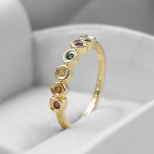 May include: A gold ring with a series of colorful gemstones. The ring features a row of round-cut stones in various colors, including red, orange, yellow, green, and purple. The ring is set in a polished gold band.