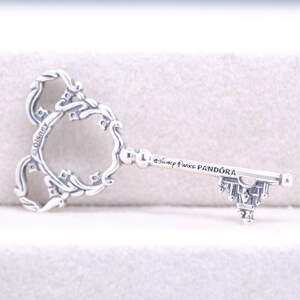 May include: A silver-colored key pendant with a decorative design. The key features a Mickey Mouse head shape and the text "Disney Parks Pandora". The key's handle has a castle-like design.