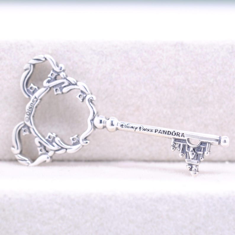 May include: A silver-colored key pendant with a decorative design. The key features a Mickey Mouse head shape and the text "Disney Parks Pandora". The key's handle has a castle-like design.