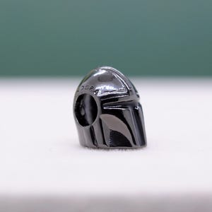 Puede incluir: Un charm de metal gris oscuro con forma de casco mandaloriano. El casco tiene un acabado brillante y presenta l&iacute;neas detalladas y una abertura circular. La palabra "DGR" es visible en el lateral del casco. El charm es probablemente para una pulsera o collar.