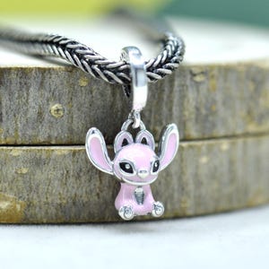 May include: A silver charm pendant featuring a pink cartoon character with large ears and black eyes. The charm is attached to a silver chain. The background is a blurred wooden surface.