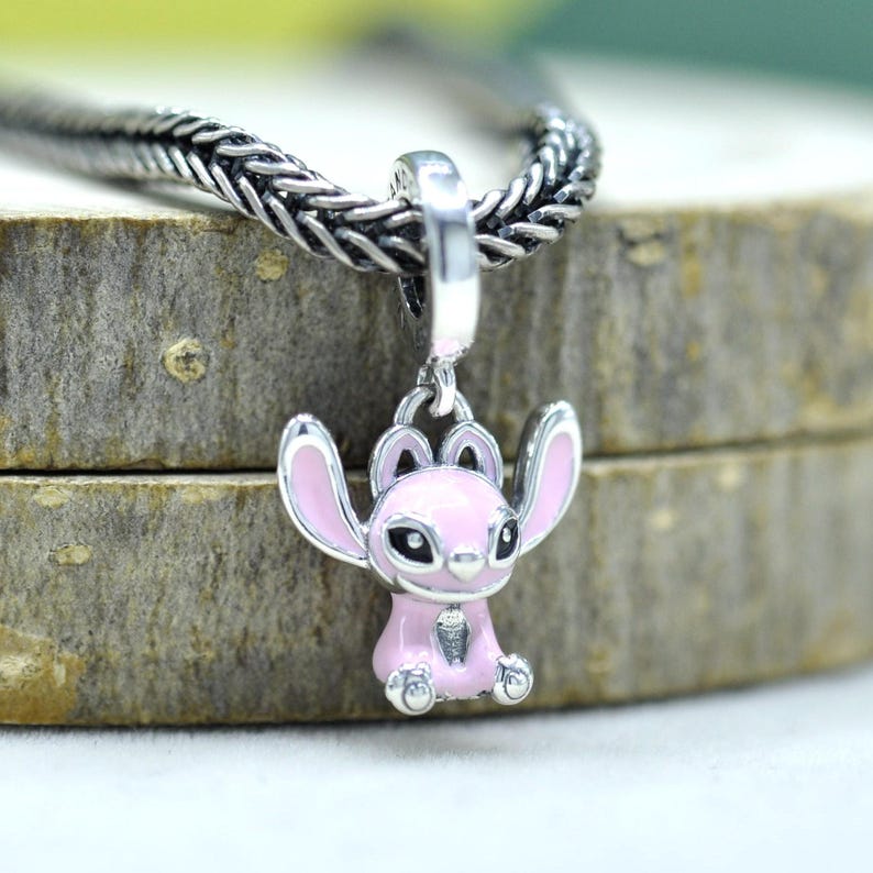 May include: A silver charm pendant featuring a pink cartoon character with large ears and black eyes. The charm is attached to a silver chain. The background is a blurred wooden surface.