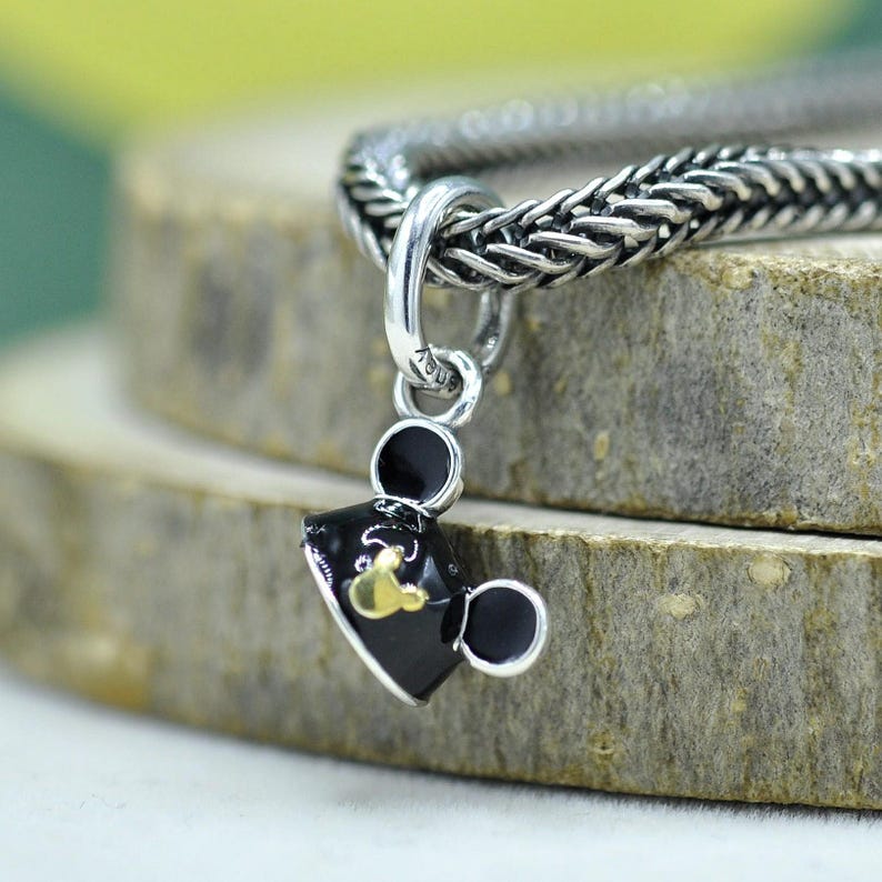 May include: A silver charm with a black and gold Mickey Mouse head design. The charm is hanging from a silver chain. The charm is a small, detailed piece of jewelry.