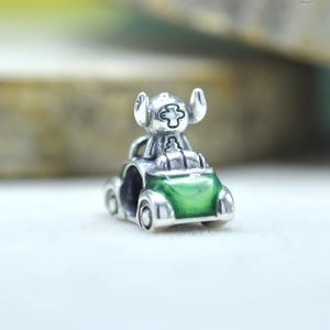 May include: A silver charm bead featuring a small creature with large ears sitting inside a green enameled car. The character has a cross-shaped marking on its forehead and is positioned in the driver seat of the metallic vehicle with wheels.