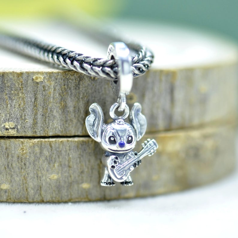 May include: A silver charm shaped like the character Stitch from Lilo and Stitch, featuring large ears and holding a small guitar. The charm is suspended from a silver chain, with a blue enamel nose and black eyes, crafted with detailed metal textures.