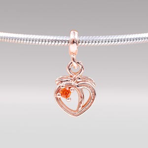 May include: A rose gold charm in the shape of a peach, with a small orange gemstone in the center. The charm is suspended from a silver chain. The peach design has a detailed outline and leaf accents.