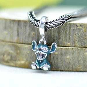 May include: A silver charm featuring a teal-colored cartoon character with large ears and a wide grin. The charm is suspended from a silver chain. The character's eyes are closed, and its mouth is open, revealing teeth. The charm is approximately 1 inch in height.
