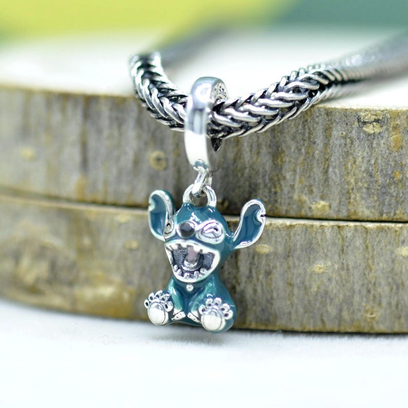 May include: A silver charm featuring a teal-colored cartoon character with large ears and a wide grin. The charm is suspended from a silver chain. The character's eyes are closed, and its mouth is open, revealing teeth. The charm is approximately 1 inch in height.