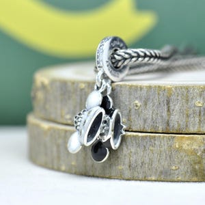 May include: A silver charm with a Mickey Mouse design. The charm features black and white enamel details, a small silver crown, and a beaded chain. The charm is displayed on a wooden surface.