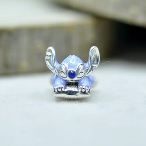 May include: A silver-toned metal charm featuring a light blue enameled character with large ears, a dark blue nose, and a smiling expression. The character is depicted in a resting position with its front paws extended forward on a silver base.