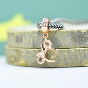 May include: A rose gold charm pendant with a floral design. The charm is attached to a braided silver ring and a silver chain. The pendant features a stylized floral design with three circular elements, each with a small, dark center.