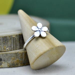 Puede incluir: Un anillo de plata con un delicado diseño floral. El anillo tiene una banda delgada y una flor de cinco pétalos con un pequeño centro redondo. El anillo se exhibe en un soporte de anillo de madera.
