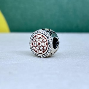 May include: A silver charm with a red and white geometric design. The charm is round with a halo of clear stones around the red center. The sides of the charm have a patterned design.