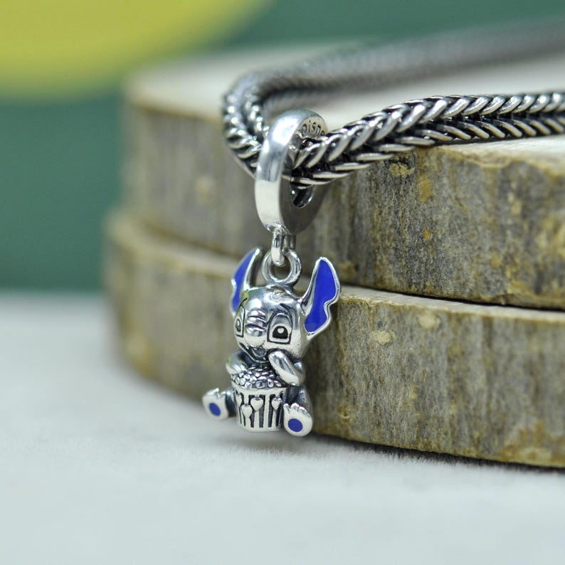 May include: A silver charm featuring the character Stitch from Lilo & Stitch, with blue enamel accents on the ears and feet. The charm is holding a cupcake and is attached to a braided silver chain.