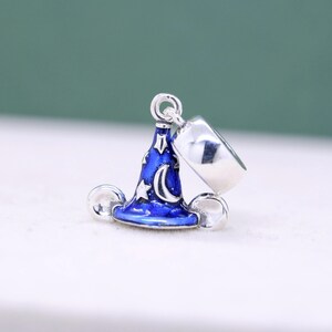 May include: A silver charm featuring a blue wizard hat with a crescent moon and stars design. The charm also includes a silver ring and two silver spheres. The hat is detailed with white accents.