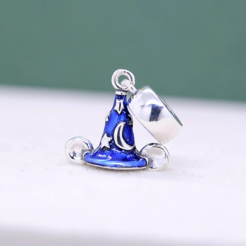 May include: A silver charm featuring a blue wizard hat with a crescent moon and stars design. The charm also includes a silver ring and two silver spheres. The hat is detailed with white accents.
