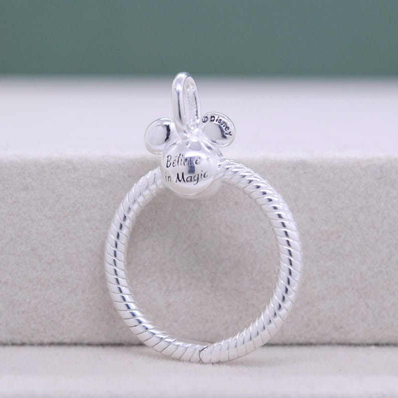 Pu&ograve; includere: Un charm argentato con un design a testa di Topolino e un anello circolare. Il charm presenta la scritta "Believe in Magic", con il logo Disney. L'anello &egrave; strutturato con un motivo a spirale.
