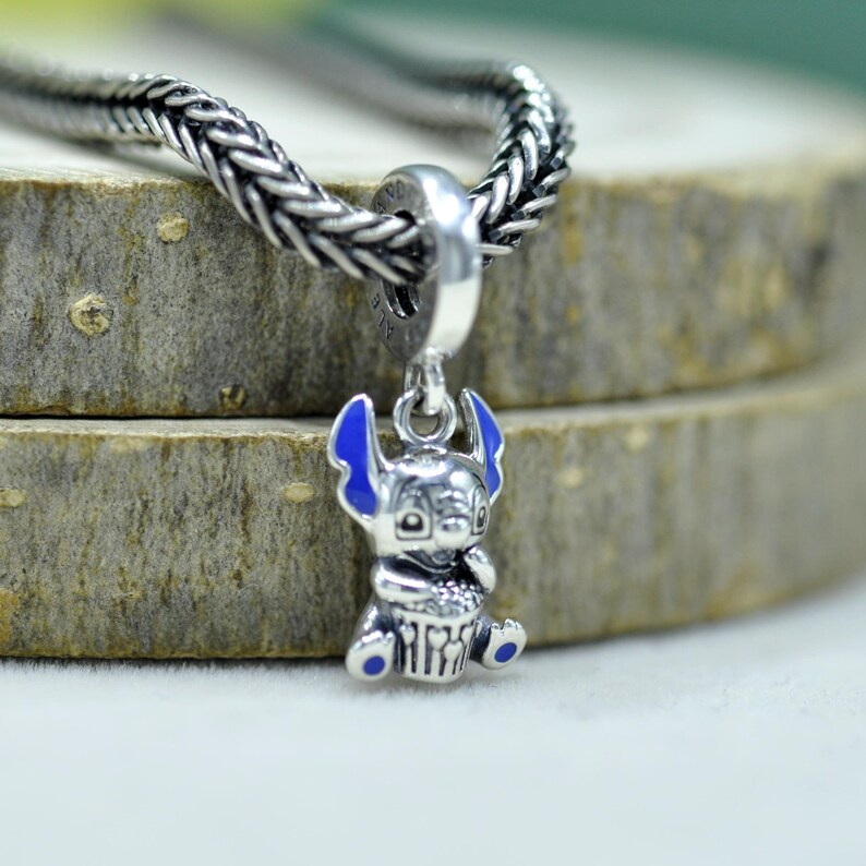 May include: A silver charm pendant featuring a cartoon character with blue accents. The charm is attached to a silver chain. The character has large ears and is holding a small object.
