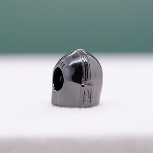 Puede incluir: Un peque&ntilde;o dije de casco gris met&aacute;lico con una parte superior redondeada y una abertura para la visera. El casco tiene una superficie lisa y reflectante y detalles grabados en el lateral. El texto "1FL" es visible.