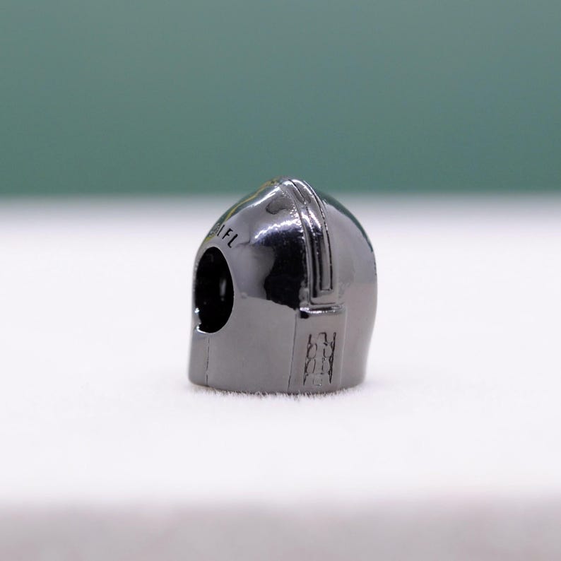 Puede incluir: Un peque&ntilde;o dije de casco gris met&aacute;lico con una parte superior redondeada y una abertura para la visera. El casco tiene una superficie lisa y reflectante y detalles grabados en el lateral. El texto "1FL" es visible.